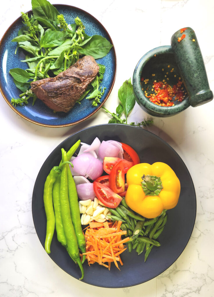 ingredients for Chilli Basil Steak, including basil and beef on a plate, vegetables and a mortar and pestle