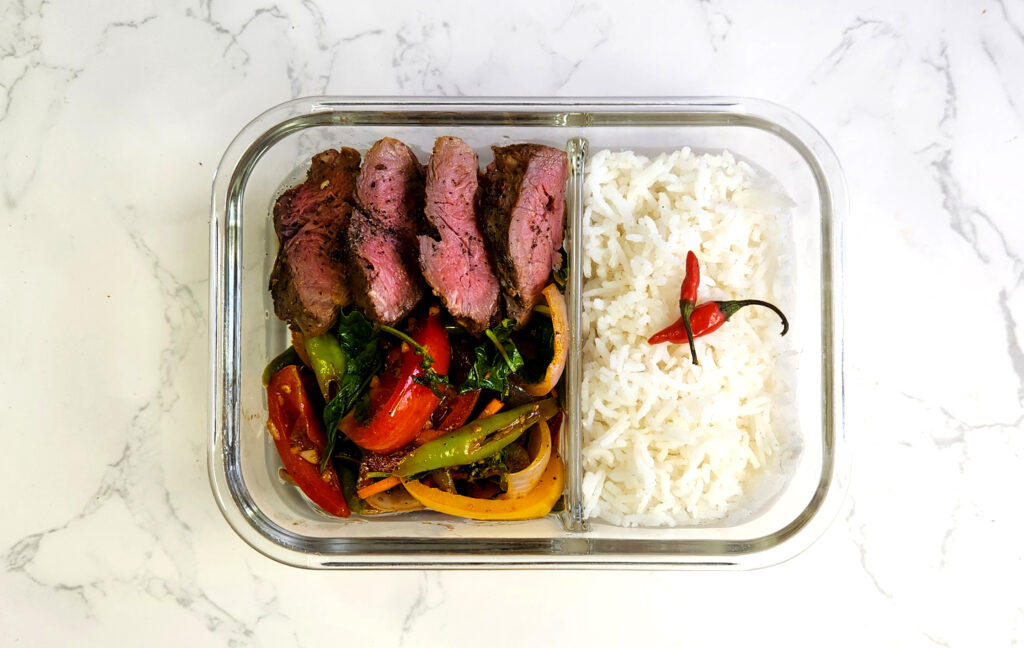 glass lunchbox with chilli basil stir fry and slices of steak and rice with chillies on it