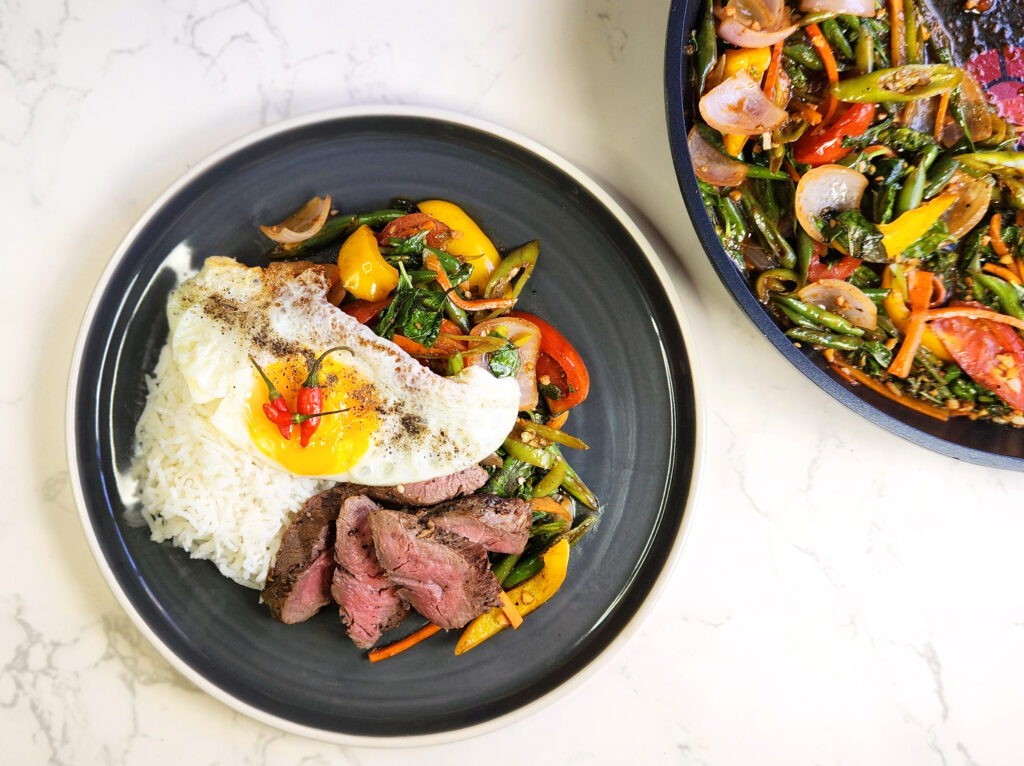 plate of chili basil steak on a bed of rice topped with an egg, stir fry pan with cooked ingredients on the side. 