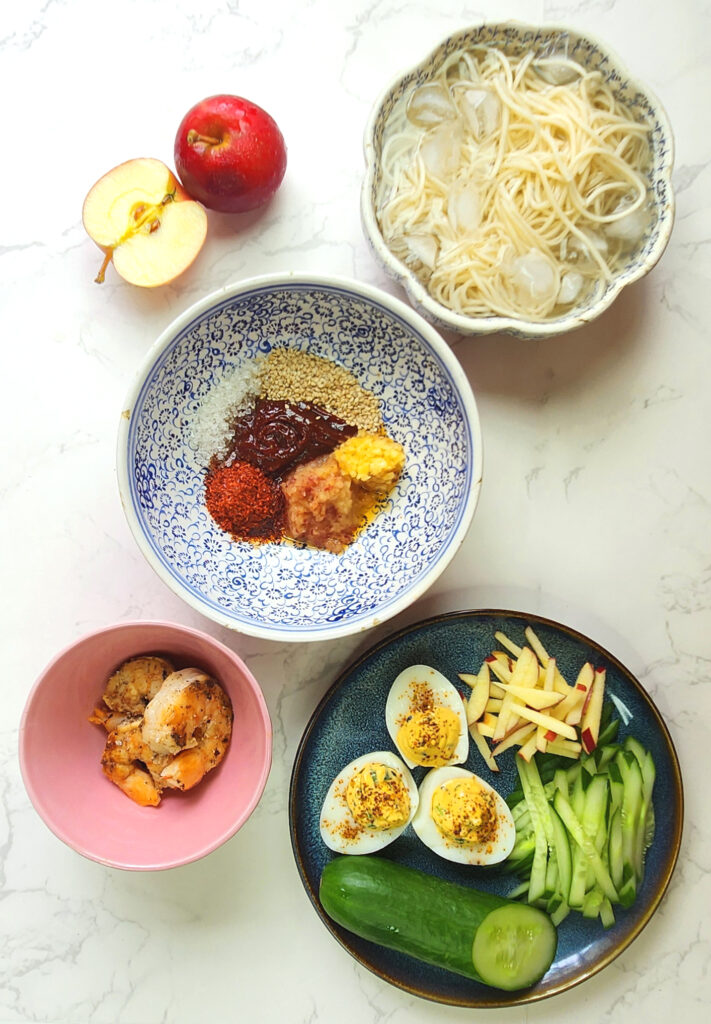 Bibimmyeon dressing ingredients in a bowl, with cold noodles, devilled eggs, cucumber and apple matchsticks and cooked prawns.
