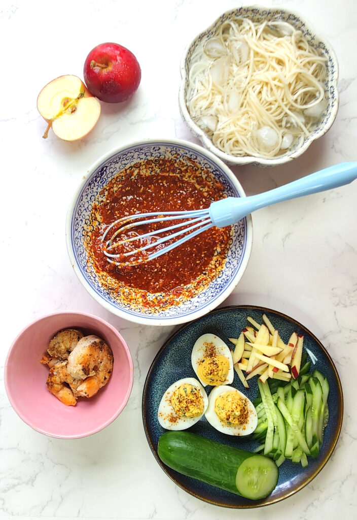 Bibimmyeon dressing ingredients combined in a bowl with a whisk, with cold noodles, devilled eggs, cucumber and apple matchsticks and cooked prawns.