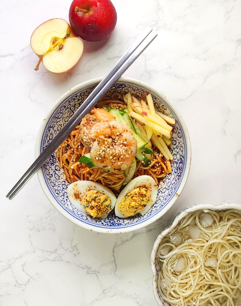 Bibimmyeon Korean cold noodles in a bowl with chopsticks, devilled eggs, cucumber, apple slices and prawn. Also whole and cut apples, noodles in ice water