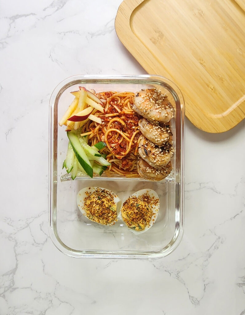 Bibimmyeon cold noodles, with cooked prawn and gochujang devilled eggs, matchsticks of cucumber and apples in a glass lunchbox