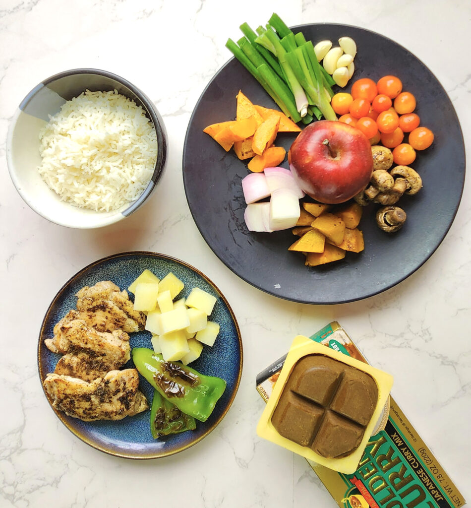 Japanese curry ingredients including steamed rice in a bowl, apple, onions, tomatoes, garlic, pumpkin and carrots, mushrooms and potatoes, and cubes of instant Japanese curry roux