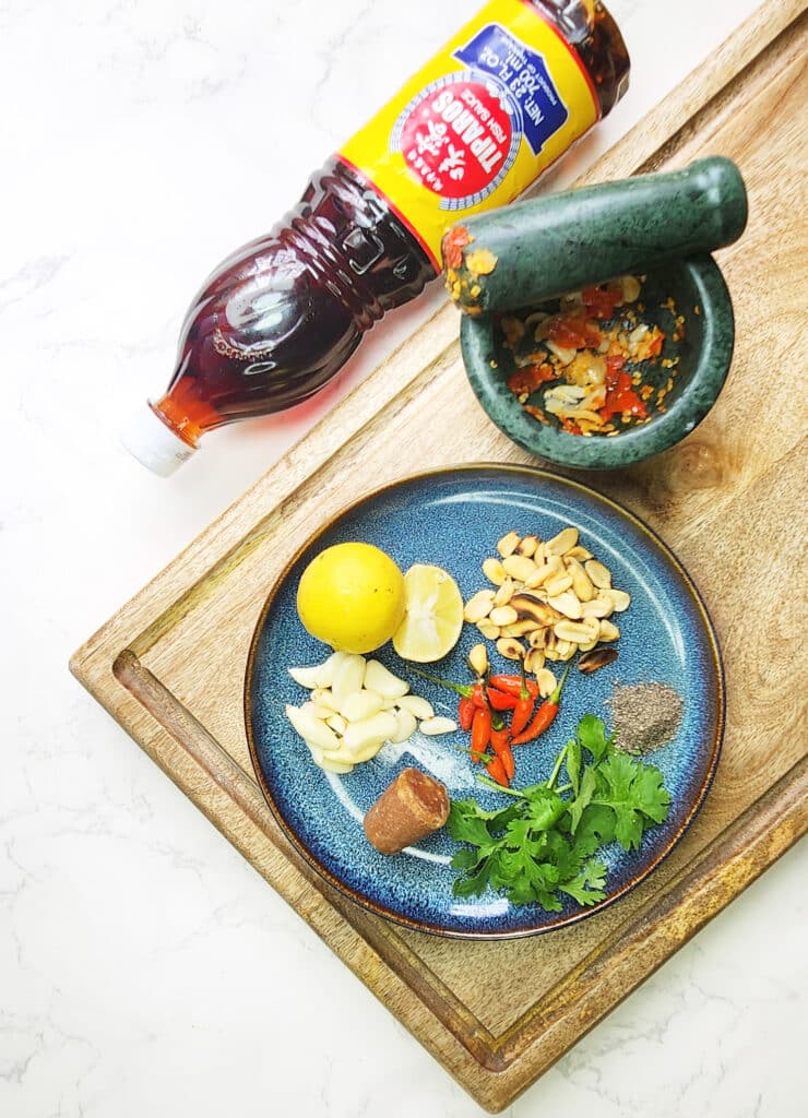 Thai Nam Jim Sauce ingredients in a mortar with a pestle, and a plate of ingredients including coriander, birds eye chilli and lime, on a wooden chopping board. Bottle of Fish Sauce. 