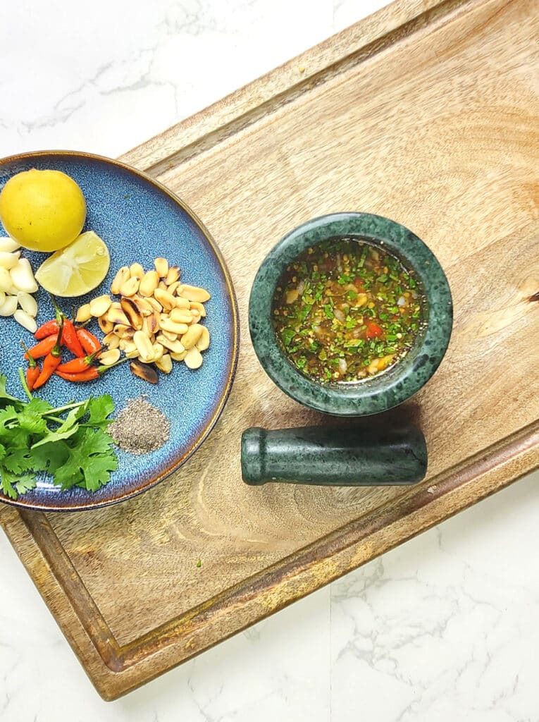 Thai Nam Jim Sauce in a mortar with a pestle, and a plate of ingredients including coriander, birds eye chilli and lime, on a wooden chopping board