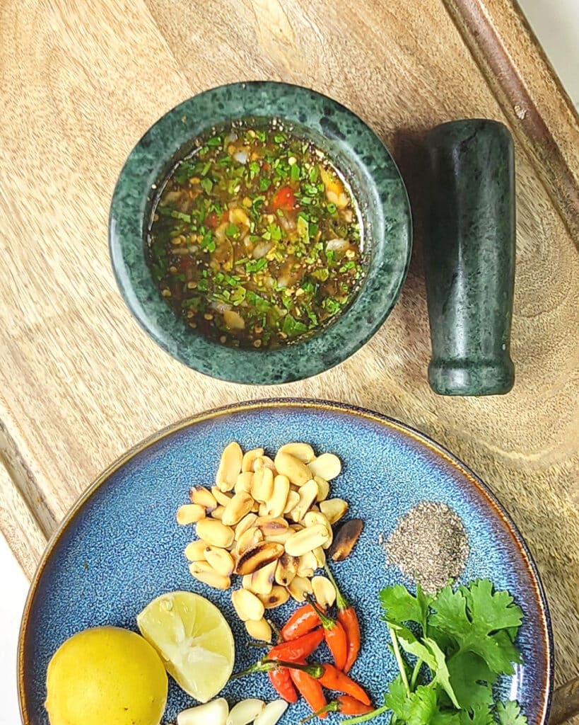 Thai Nam Jim Sauce in a mortar with a pestle, and a plate of ingredients including coriander, birds eye chilli and lime, on a wooden chopping board