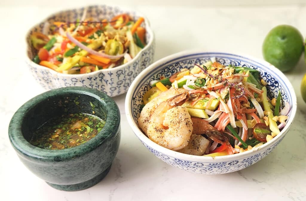 Thai Nam Jim Sauce in a stone bowl, and bowls of Thai Green Mango Salad with prawns. 