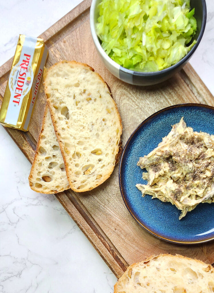 Butter, slices of sourdough bread and chicken mayonnaise sandwich filling, shredded lettuce leaves in the background.