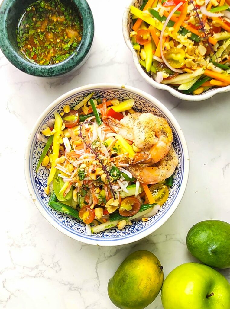 Thai Green Mango Salad with peanuts, prawns, crabsticks and Nam Jim Dressing in a bowl. Whole apples and green mangoes on the side.