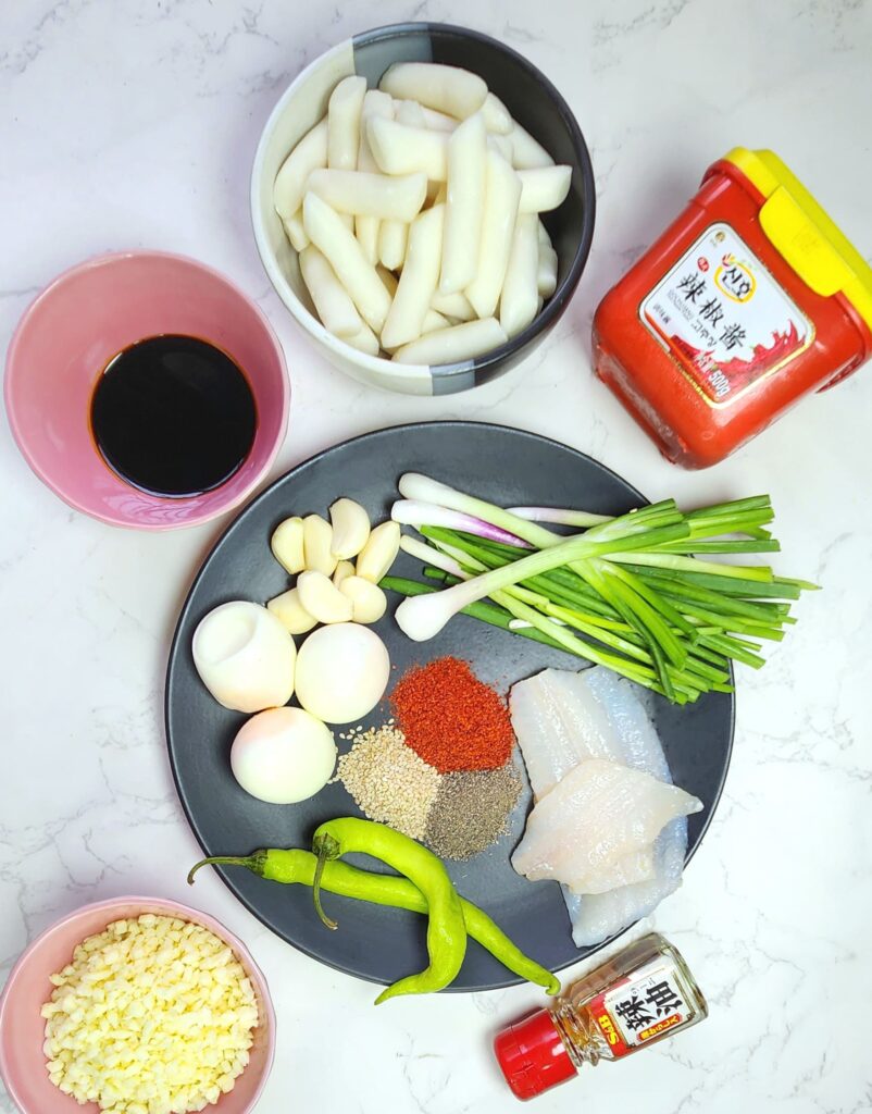 Tteokbokki ingredients Korean rice cakes in a bowl, gochujang paste, soy sauce, spring onions, garlic, hardboiled eggs, chilli powder, fish fillets, sesame seeds, green chillis and shredded cheese