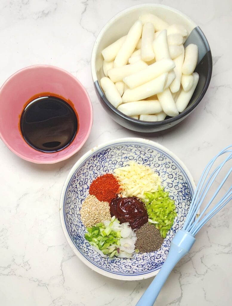 Tteokbokki ingredients Korean rice cakes in a bowl, gochujang paste, soy sauce, spring onions, garlic, chilli powder, sesame seeds, green chillis and pepper