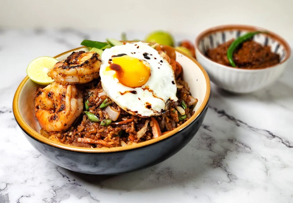 Nasi Goreng - Indonesian fried rice in a bowl with prawns, cherry tomatoes, cucumbers, lime and a fried egg, sambal terasi