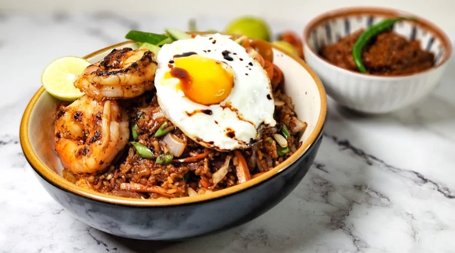 Nasi Goreng Indonesian fried rice in a bowl with prawns, cherry tomatoes, cucumbers, lime and a fried egg, sambal terasi