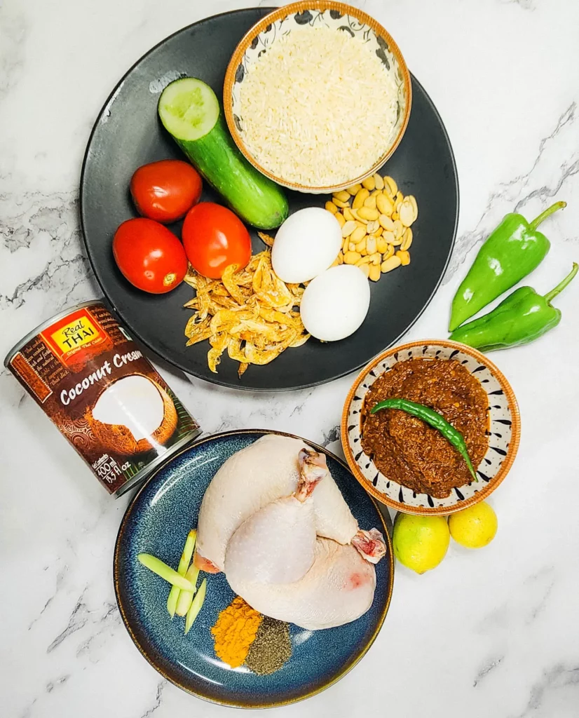 Nasi Lemak ingredients on a plate - coconut cream, rice, chillies, dried shrimp, eggs, sambal and peanuts, lemongrass, chicken, turmeric and pepper, lime and tomato and cucumber