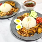 Nasi Lemak on a plate - coconut rice with dried shrimp, boiled eggs, sambal and peanuts, lemongrass grilled chicken and tomato and cucumber slices