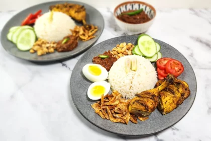 Nasi Lemak on a plate - coconut rice with dried shrimp, boiled eggs, sambal and peanuts, lemongrass grilled chicken and tomato and cucumber slices