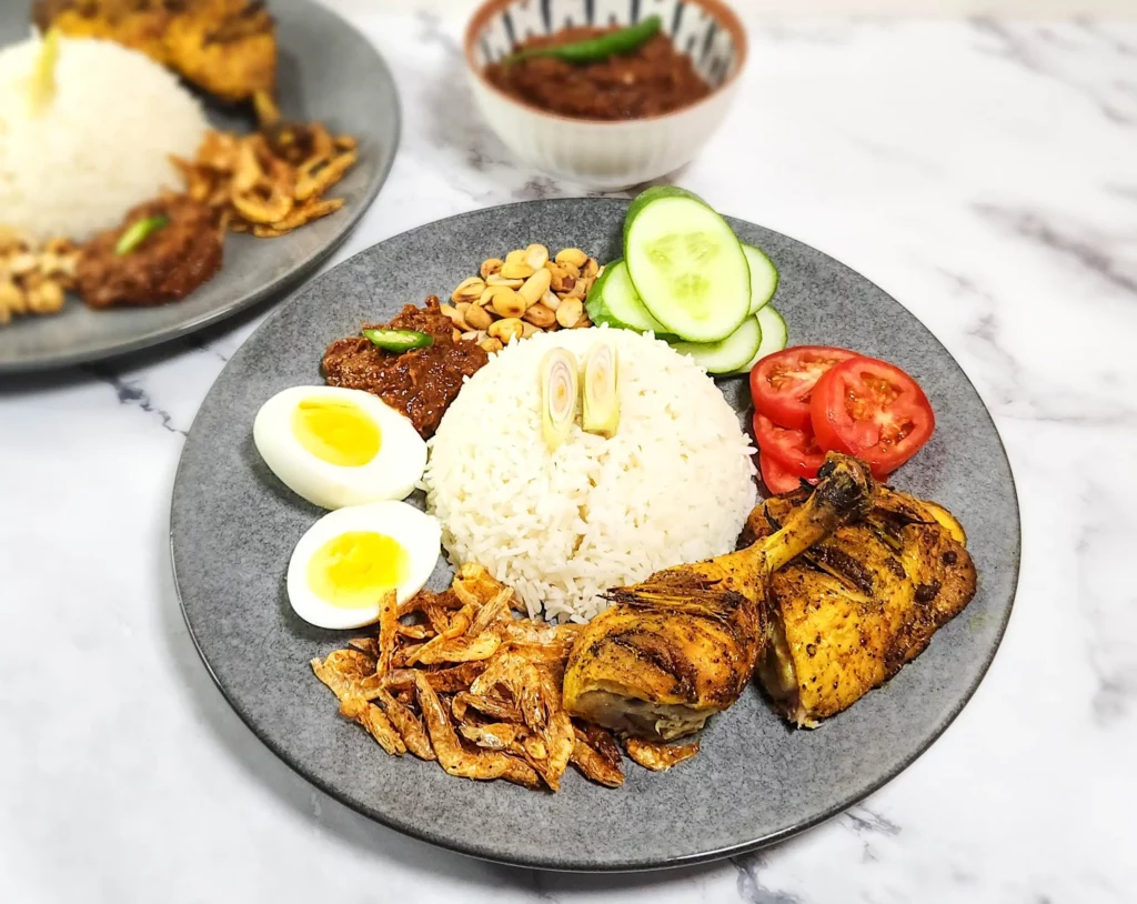 Nasi Lemak on a plate - coconut rice with dried shrimp, boiled eggs, sambal and peanuts, lemongrass grilled chicken and tomato and cucumber slices