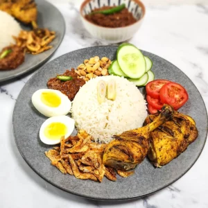 Nasi Lemak on a plate - coconut rice with dried shrimp, boiled eggs, sambal and peanuts, lemongrass grilled chicken and tomato and cucumber slices