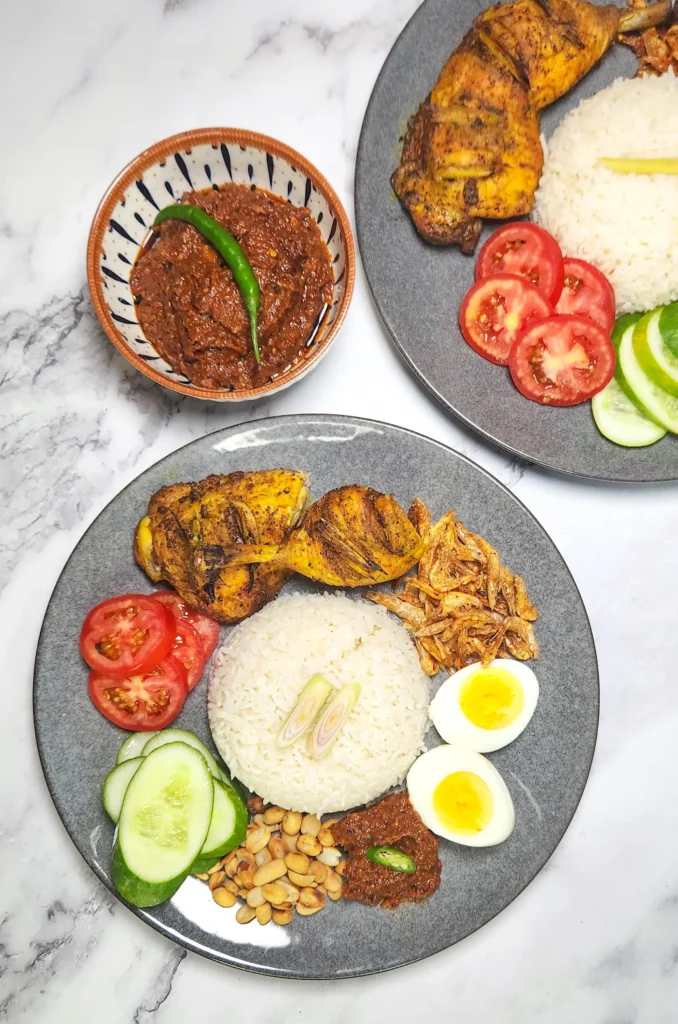 Nasi Lemak on a plate - coconut rice with dried shrimp, boiled eggs, sambal and peanuts, lemongrass grilled chicken and tomato and cucumber slices