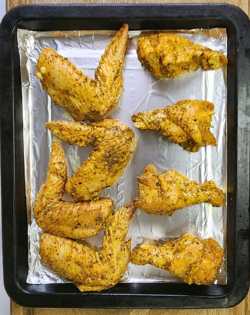 dry rub chicken wings ingredients for mix including pepper, chilli, garlic powder, salt sprinkled on chicken wings laid on baking tray